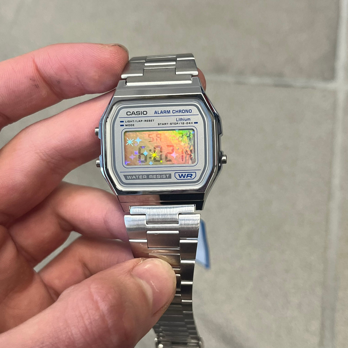Custom Casio A158 "Double Holo" Silver Digital Watch (A158WEA-7EF) is a custom Casio digital watch with a Double Holo (Stars and normal holo) filter over the square resin glass screen and a stainless steel band with an adjustable clasp, compatible with wrist sizes from 150 to 205 mm. It has a 1/100-second stopwatch, a daily alarm, an hourly time signal, an auto-calendar, a 12/24-hour format, a green LED light, and water resistance.