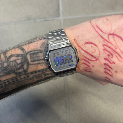 Custom Casio A158 "Double Holo" Silver Digital Watch (A158WEA-7EF) is a custom Casio digital watch with a Double Holo (Stars and normal holo) filter over the square resin glass screen and a stainless steel band with an adjustable clasp, compatible with wrist sizes from 150 to 205 mm. It has a 1/100-second stopwatch, a daily alarm, an hourly time signal, an auto-calendar, a 12/24-hour format, a green LED light, and water resistance.