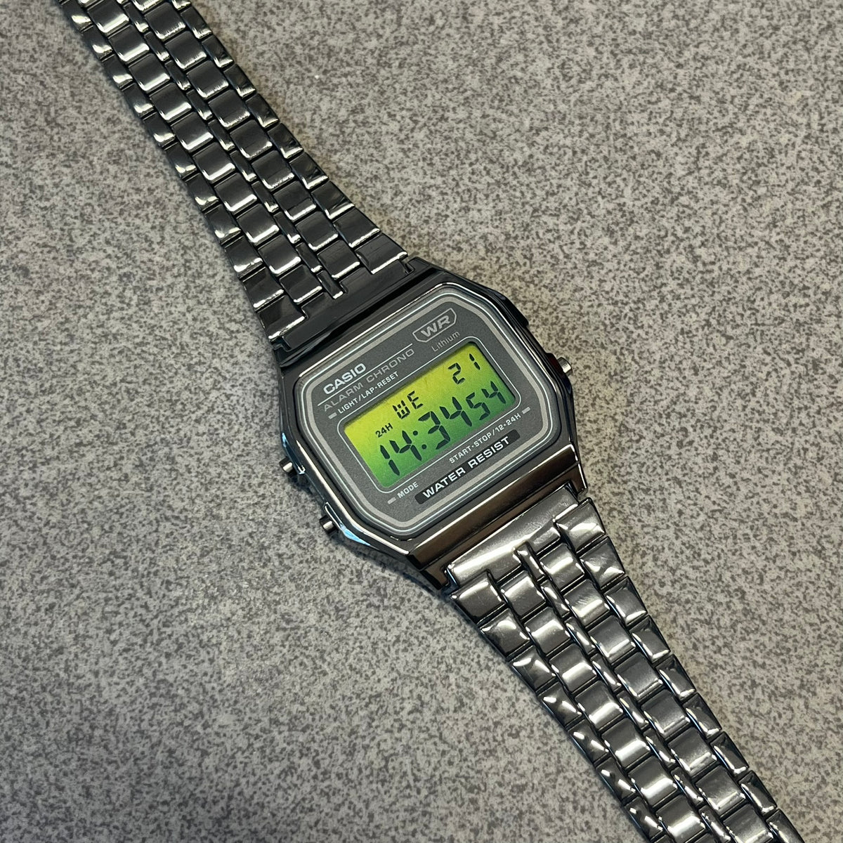 Custom Casio A158 “Emerald” Green &amp; Black Digital Watch is a watch with a green gradient filter over the square screen and a grey ion-plated stainless steel band with an adjustable clasp. The case comes with a resin glass and a resin and black chrome-plated case and bezel. It has a 1/100-second stopwatch, a daily alarm, an hourly time signal, an auto-calendar, a 12/24-hour format, a green LED light, and water resistance.