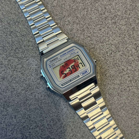 Custom Casio A158 “Kiss Me” Red &amp; Silver Digital Watch (A158WEA-7EF) is a custom Casio digital watch with a red lips filter over the square resin glass screen and a stainless steel band with an adjustable clasp, compatible with wrist sizes from 150 to 205 mm. It has a 1/100-second stopwatch, a daily alarm, an hourly time signal, an auto-calendar, a 12/24-hour format, a green LED light, and water resistance.