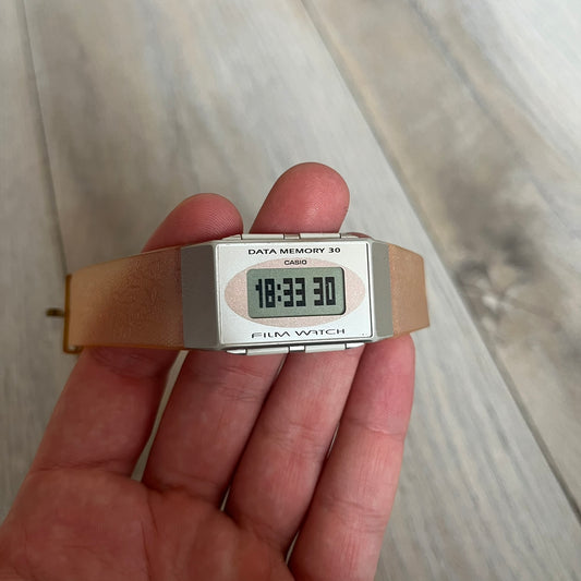Casio FS-06 Data Memory-30 “Film Watch” is a vintage mid-1980s digital timepiece featuring a rectangular silver case with salmon accents. It includes a semi-transparent orange strap with a floral pattern and a strong blue backlight display. The watch offers multiple functions such as world time, dual alarms, chronograph, and memo/data memory mode. It is made in Malaysia and features tactile buttons, illuminated display, and a signed case back.