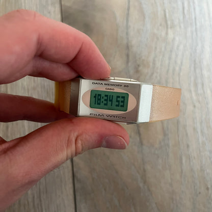 Casio FS-06 Data Memory-30 “Film Watch” is a vintage mid-1980s digital timepiece featuring a rectangular silver case with salmon accents. It includes a semi-transparent orange strap with a floral pattern and a strong blue backlight display. The watch offers multiple functions such as world time, dual alarms, chronograph, and memo/data memory mode. It is made in Malaysia and features tactile buttons, illuminated display, and a signed case back.