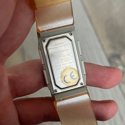 Casio FS-06 Data Memory-30 “Film Watch” is a vintage mid-1980s digital timepiece featuring a rectangular silver case with salmon accents. It includes a semi-transparent orange strap with a floral pattern and a strong blue backlight display. The watch offers multiple functions such as world time, dual alarms, chronograph, and memo/data memory mode. It is made in Malaysia and features tactile buttons, illuminated display, and a signed case back.
