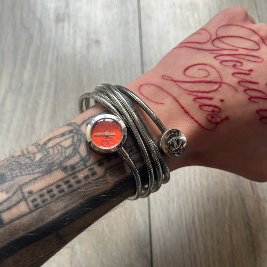 Just Cavalli 2000’s Red Stars Bracelet Watch is a stretch metal bracelet watch from Just Cavalli featuring multiple intersecting silver-tone coil bands that create a layered wrap-around design. One end of the bracelet holds a small round analog watch with a red dial finished with a textured star pattern, while the opposite end features a circular metal element engraved with the JC logo. The bracelet uses a spring-style stretch construction instead of a clasp, allowing it to expand to fit the wrist. The watc