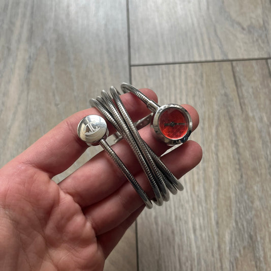 Just Cavalli 2000’s Red Stars Bracelet Watch is a stretch metal bracelet watch from Just Cavalli featuring multiple intersecting silver-tone coil bands that create a layered wrap-around design. One end of the bracelet holds a small round analog watch with a red dial finished with a textured star pattern, while the opposite end features a circular metal element engraved with the JC logo. The bracelet uses a spring-style stretch construction instead of a clasp, allowing it to expand to fit the wrist. The watc