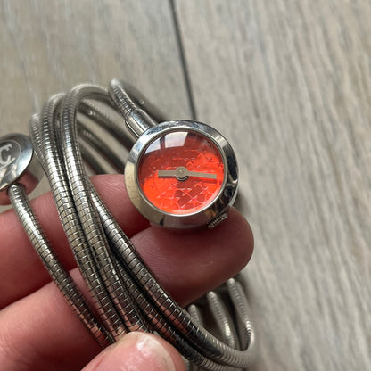 Just Cavalli 2000’s Red Stars Bracelet Watch is a stretch metal bracelet watch from Just Cavalli featuring multiple intersecting silver-tone coil bands that create a layered wrap-around design. One end of the bracelet holds a small round analog watch with a red dial finished with a textured star pattern, while the opposite end features a circular metal element engraved with the JC logo. The bracelet uses a spring-style stretch construction instead of a clasp, allowing it to expand to fit the wrist. The watc