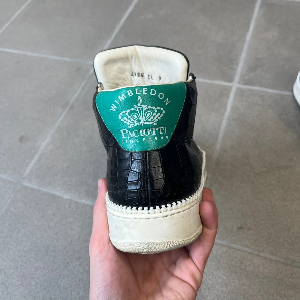 Cesare Paciotti Wimbledon Cocco Lux Black Sneakers (CPM5121) are made in Italy from black crocodile-embossed leather, with a Paciotti Wimbledon tag showing two rackets on the tongue and a second logo on the heel. The white rubber sole has a textured zipper-teeth detail running around its edge. They come with the original box, which shows wear.