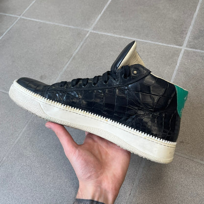 Cesare Paciotti Wimbledon Cocco Lux Black Sneakers (CPM5121) are made in Italy from black crocodile-embossed leather, with a Paciotti Wimbledon tag showing two rackets on the tongue and a second logo on the heel. The white rubber sole has a textured zipper-teeth detail running around its edge. They come with the original box, which shows wear.