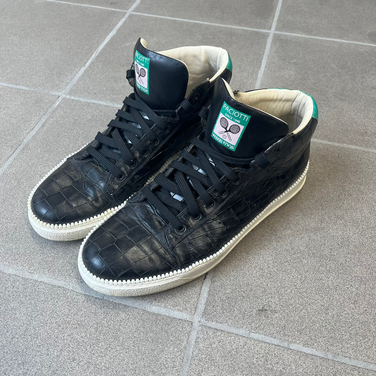 Cesare Paciotti Wimbledon Cocco Lux Black Sneakers (CPM5121) are made in Italy from black crocodile-embossed leather, with a Paciotti Wimbledon tag showing two rackets on the tongue and a second logo on the heel. The white rubber sole has a textured zipper-teeth detail running around its edge. They come with the original box, which shows wear.