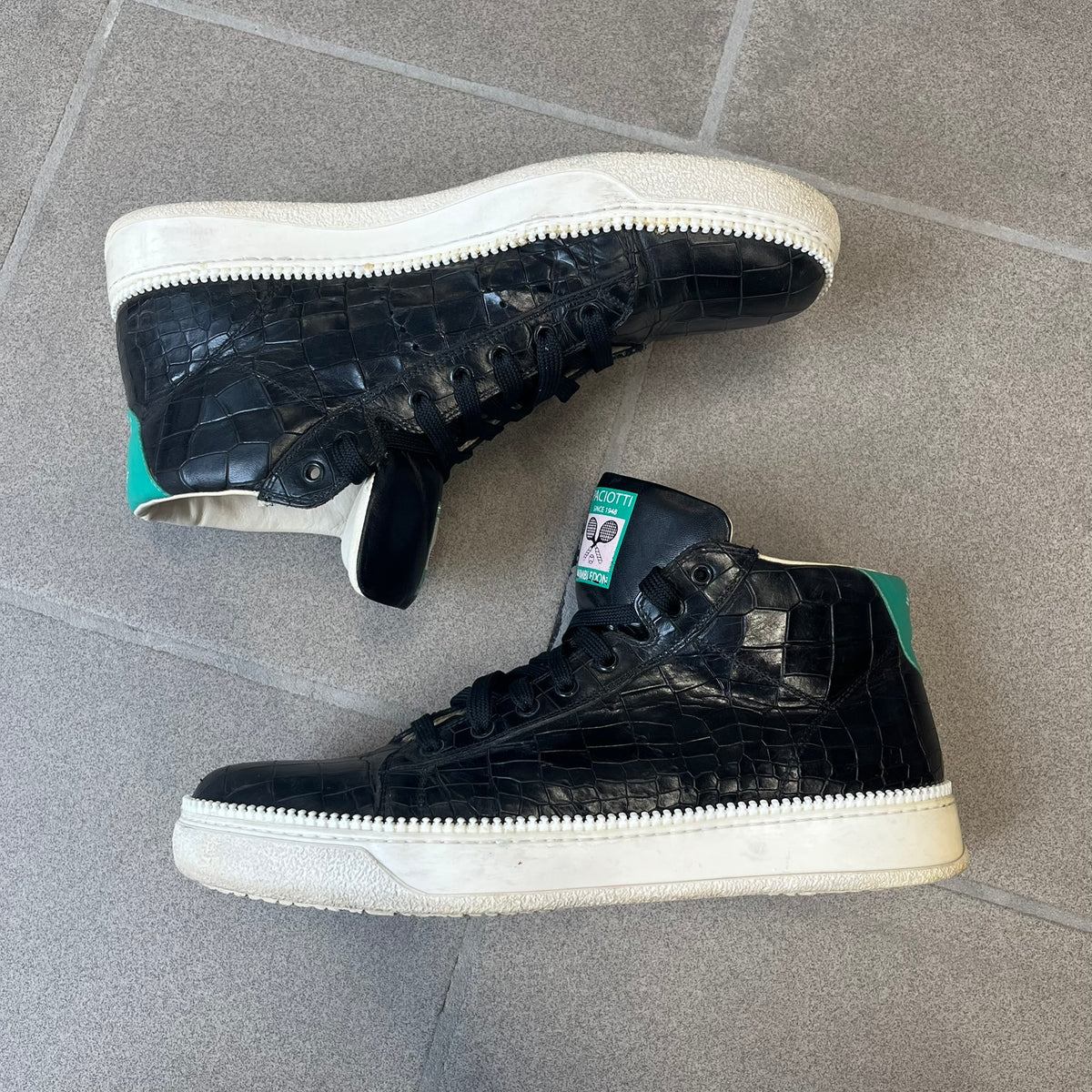 Cesare Paciotti Wimbledon Cocco Lux Black Sneakers (CPM5121) are made in Italy from black crocodile-embossed leather, with a Paciotti Wimbledon tag showing two rackets on the tongue and a second logo on the heel. The white rubber sole has a textured zipper-teeth detail running around its edge. They come with the original box, which shows wear.