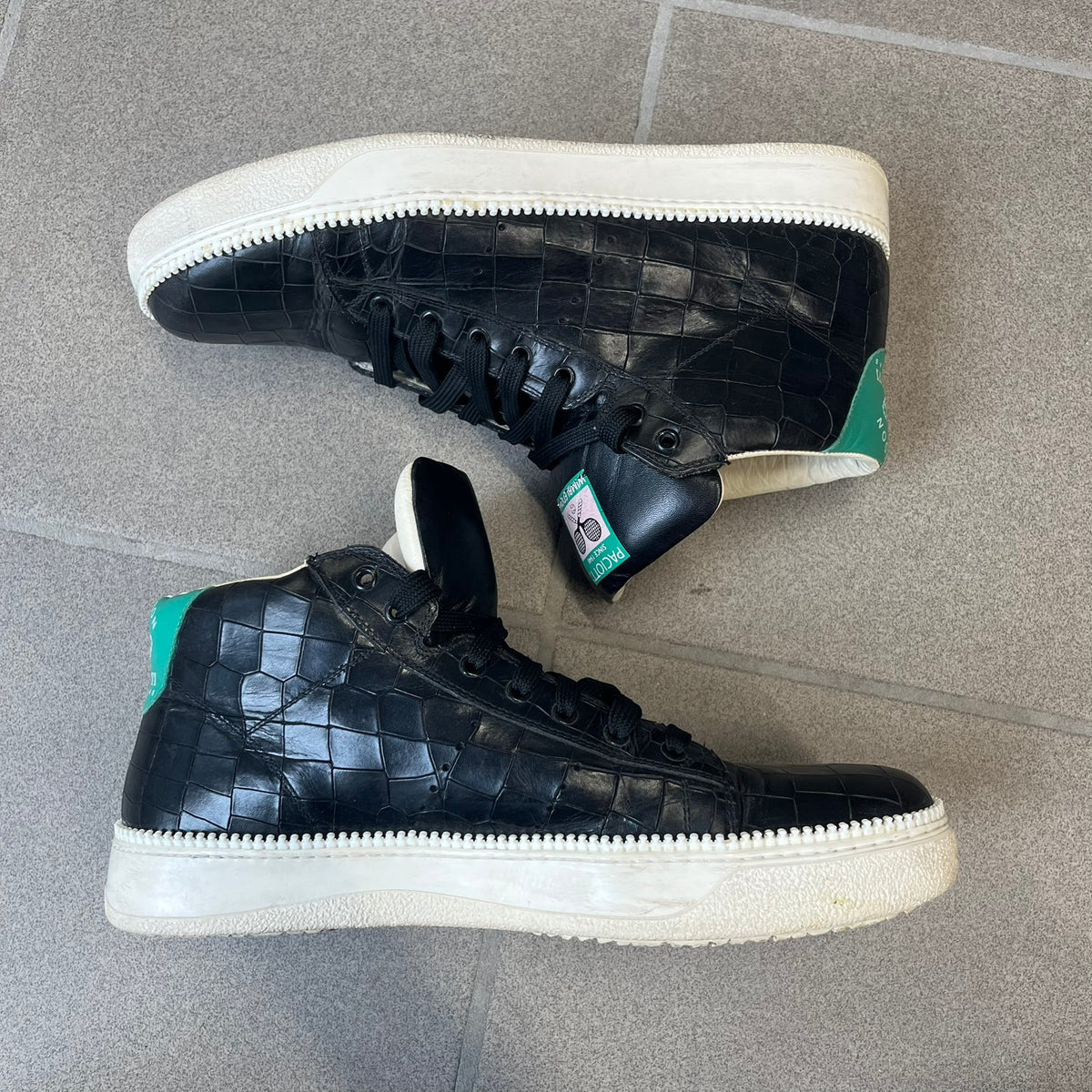 Cesare Paciotti Wimbledon Cocco Lux Black Sneakers (CPM5121) are made in Italy from black crocodile-embossed leather, with a Paciotti Wimbledon tag showing two rackets on the tongue and a second logo on the heel. The white rubber sole has a textured zipper-teeth detail running around its edge. They come with the original box, which shows wear.