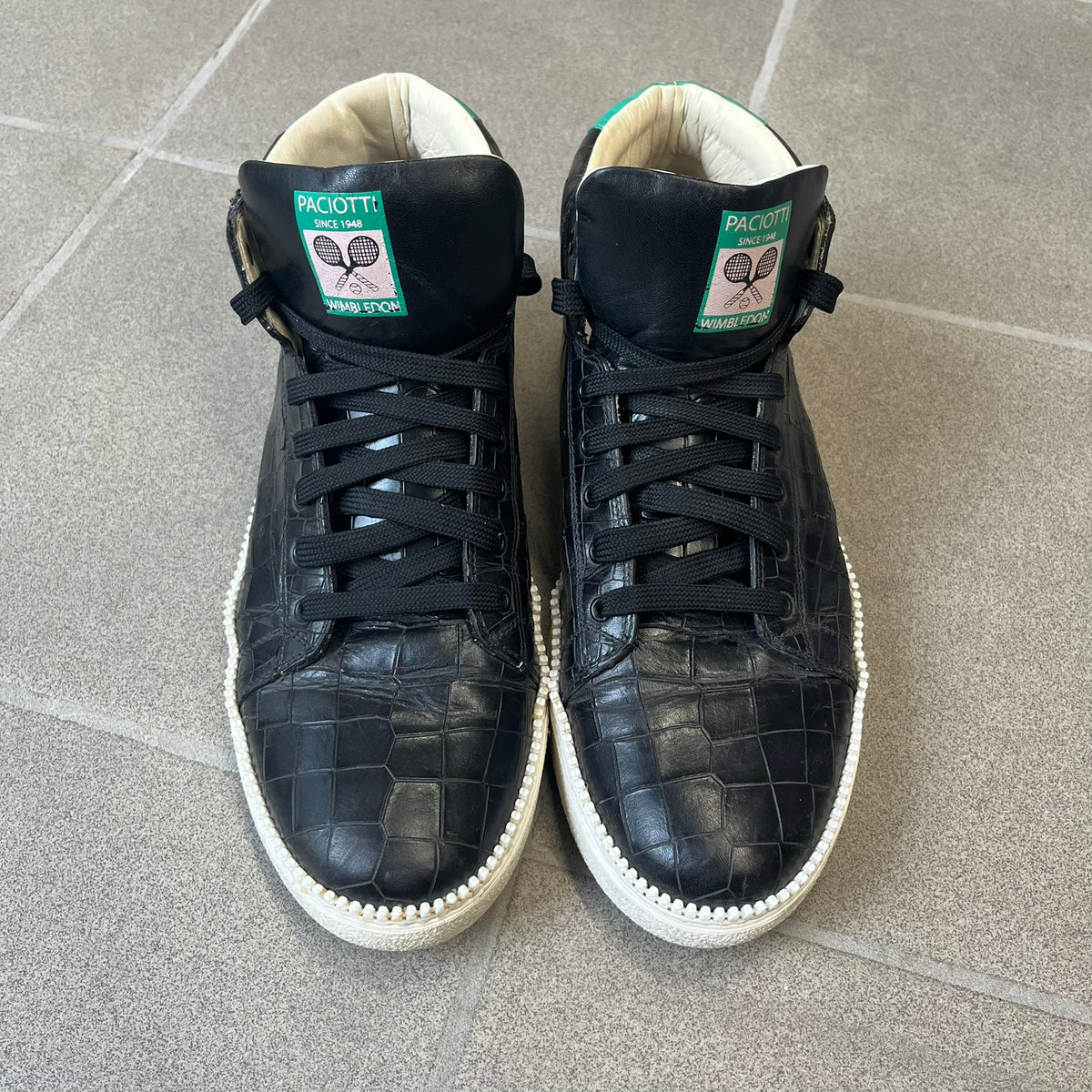 Cesare Paciotti Wimbledon Cocco Lux Black Sneakers (CPM5121) are made in Italy from black crocodile-embossed leather, with a Paciotti Wimbledon tag showing two rackets on the tongue and a second logo on the heel. The white rubber sole has a textured zipper-teeth detail running around its edge. They come with the original box, which shows wear.