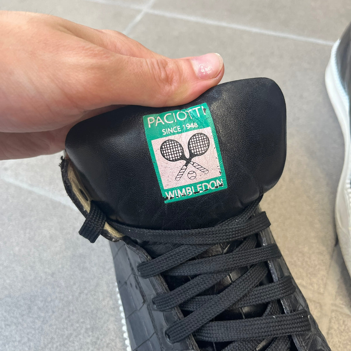 Cesare Paciotti Wimbledon Cocco Lux Black Sneakers (CPM5121) are made in Italy from black crocodile-embossed leather, with a Paciotti Wimbledon tag showing two rackets on the tongue and a second logo on the heel. The white rubber sole has a textured zipper-teeth detail running around its edge. They come with the original box, which shows wear.