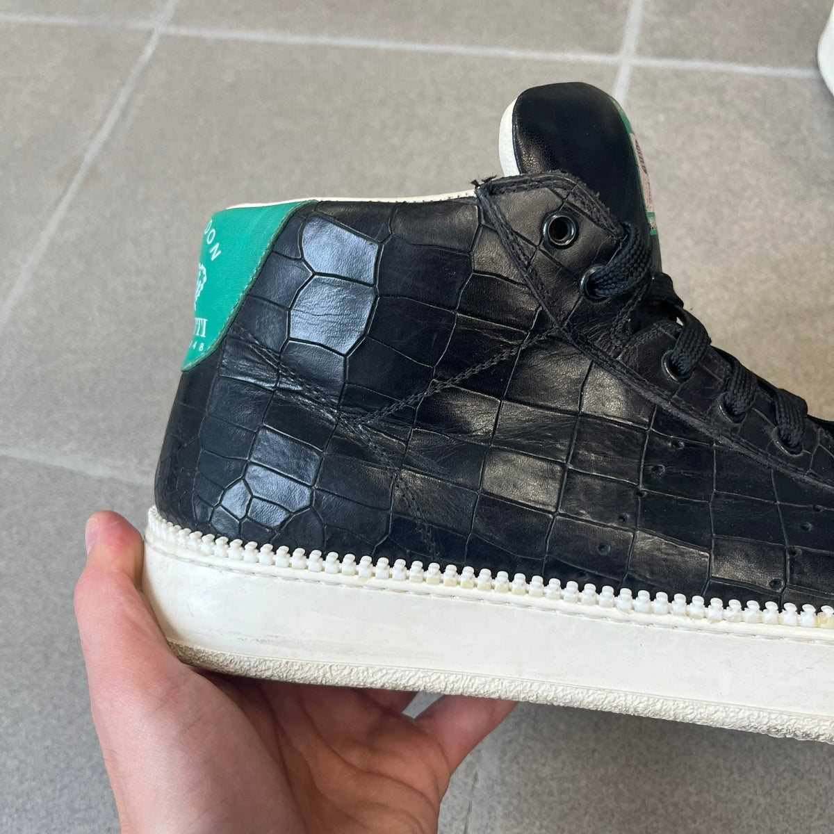 Cesare Paciotti Wimbledon Cocco Lux Black Sneakers (CPM5121) are made in Italy from black crocodile-embossed leather, with a Paciotti Wimbledon tag showing two rackets on the tongue and a second logo on the heel. The white rubber sole has a textured zipper-teeth detail running around its edge. They come with the original box, which shows wear.