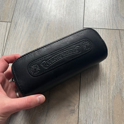 The Chrome Hearts Black Leather Sunglasses Case is a black leather zip case with a Chrome Hearts logo on the front, flanked by two cross motifs. It features a CH dagger zipper pull made from black plastic and metal. The interior is monogrammed with vine dagger logos and includes an attached soft inner compartment designed to hold a cleaning cloth.