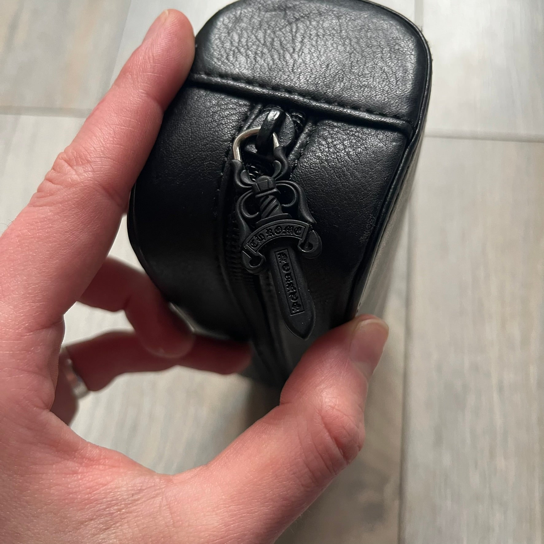 The Chrome Hearts Black Leather Sunglasses Case is a black leather zip case with a Chrome Hearts logo on the front, flanked by two cross motifs. It features a CH dagger zipper pull made from black plastic and metal. The interior is monogrammed with vine dagger logos and includes an attached soft inner compartment designed to hold a cleaning cloth.