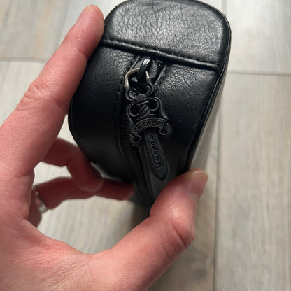 The Chrome Hearts Black Leather Sunglasses Case is a black leather zip case with a Chrome Hearts logo on the front, flanked by two cross motifs. It features a CH dagger zipper pull made from black plastic and metal. The interior is monogrammed with vine dagger logos and includes an attached soft inner compartment designed to hold a cleaning cloth.
