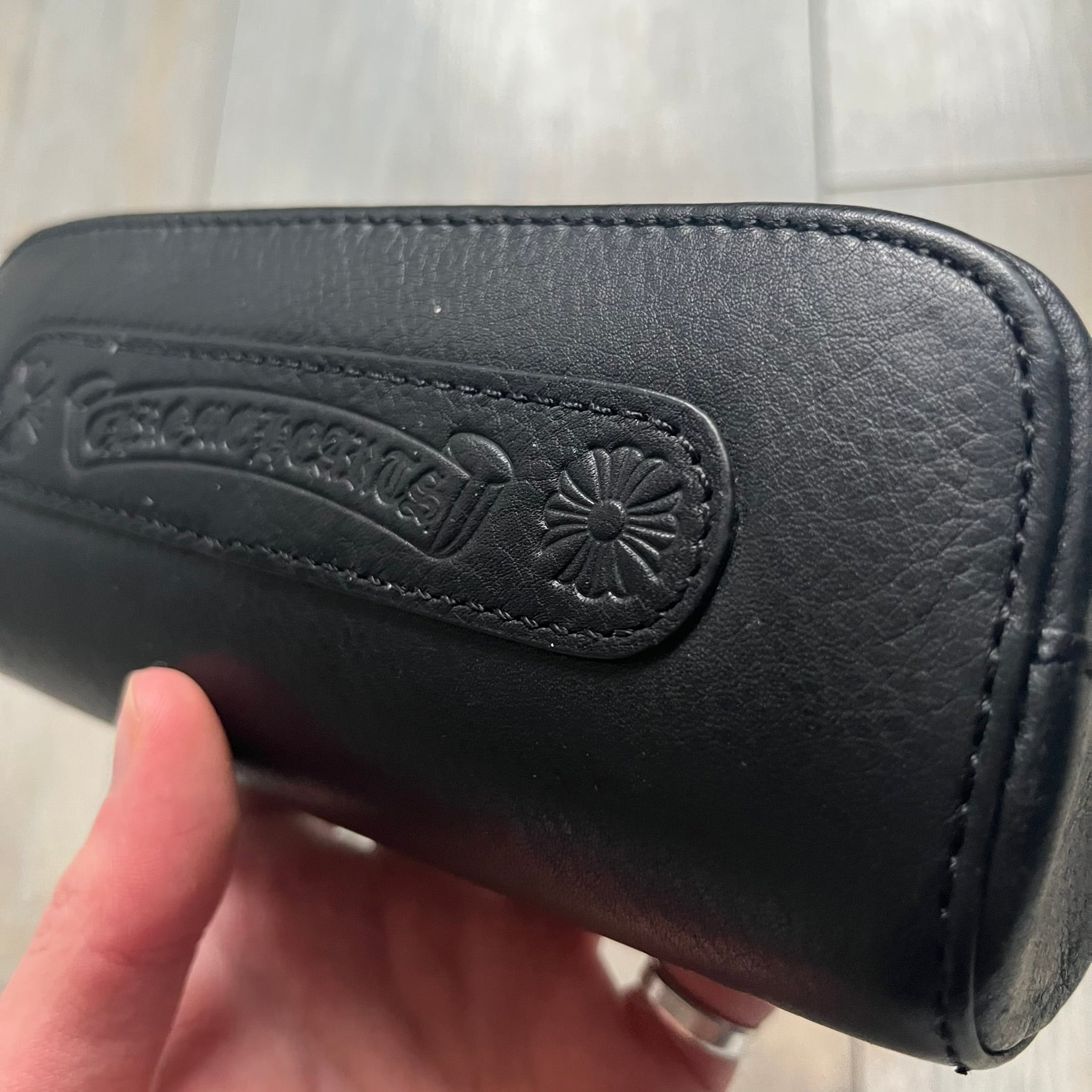 The Chrome Hearts Black Leather Sunglasses Case is a black leather zip case with a Chrome Hearts logo on the front, flanked by two cross motifs. It features a CH dagger zipper pull made from black plastic and metal. The interior is monogrammed with vine dagger logos and includes an attached soft inner compartment designed to hold a cleaning cloth.