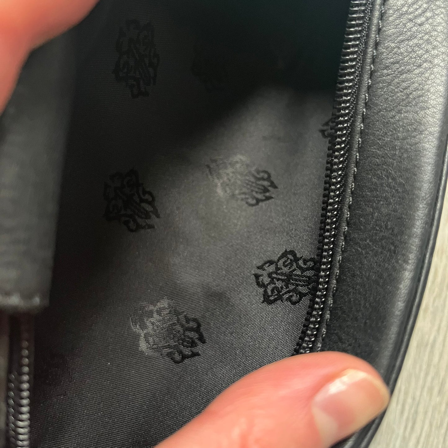 The Chrome Hearts Black Leather Sunglasses Case is a black leather zip case with a Chrome Hearts logo on the front, flanked by two cross motifs. It features a CH dagger zipper pull made from black plastic and metal. The interior is monogrammed with vine dagger logos and includes an attached soft inner compartment designed to hold a cleaning cloth.