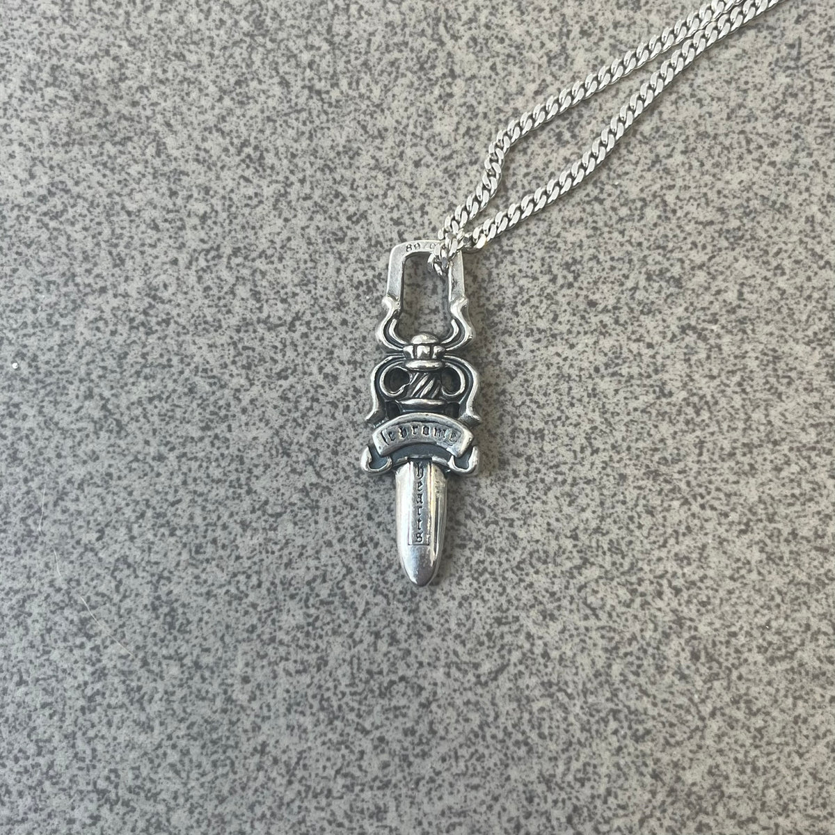 Chrome Hearts Dagger Zipper Pendant crafted from .925 silver, featuring the brand’s signature double-sided dagger design with “Chrome” on the guard and “Hearts” on the blade. Comes on a standard 925 Silver chain (non-CH). Square bail for chain attachment with 925 tag on one side and 89 03 on the other. The dagger was originally a zipper pull.