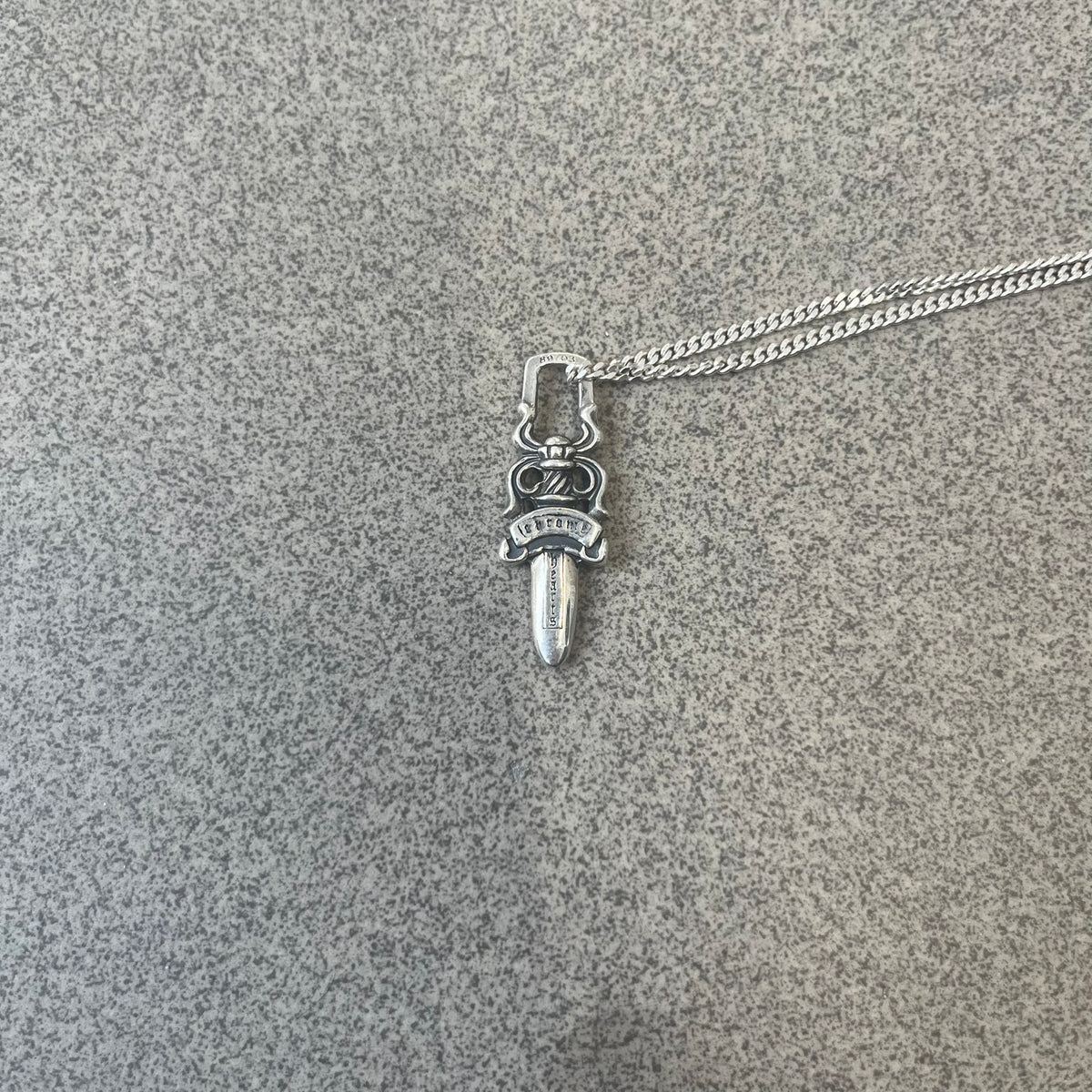 Chrome Hearts Dagger Zipper Pendant crafted from .925 silver, featuring the brand’s signature double-sided dagger design with “Chrome” on the guard and “Hearts” on the blade. Comes on a standard 925 Silver chain (non-CH). Square bail for chain attachment with 925 tag on one side and 89 03 on the other. The dagger was originally a zipper pull.