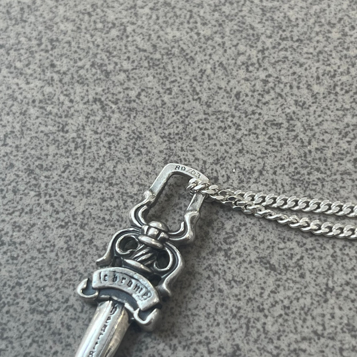 Chrome Hearts Dagger Zipper Pendant crafted from .925 silver, featuring the brand’s signature double-sided dagger design with “Chrome” on the guard and “Hearts” on the blade. Comes on a standard 925 Silver chain (non-CH). Square bail for chain attachment with 925 tag on one side and 89 03 on the other. The dagger was originally a zipper pull.