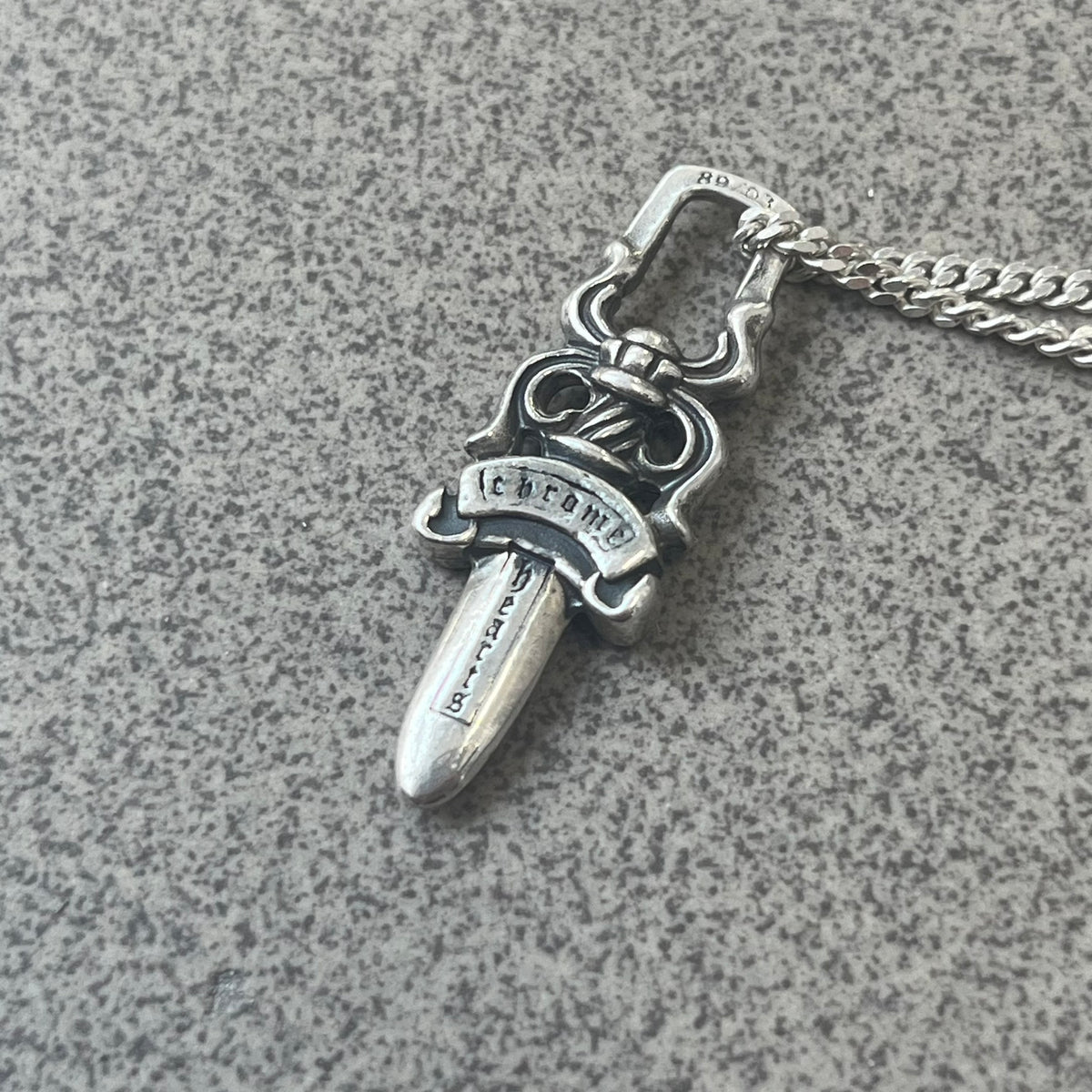 Chrome Hearts Dagger Zipper Pendant crafted from .925 silver, featuring the brand’s signature double-sided dagger design with “Chrome” on the guard and “Hearts” on the blade. Comes on a standard 925 Silver chain (non-CH). Square bail for chain attachment with 925 tag on one side and 89 03 on the other. The dagger was originally a zipper pull.