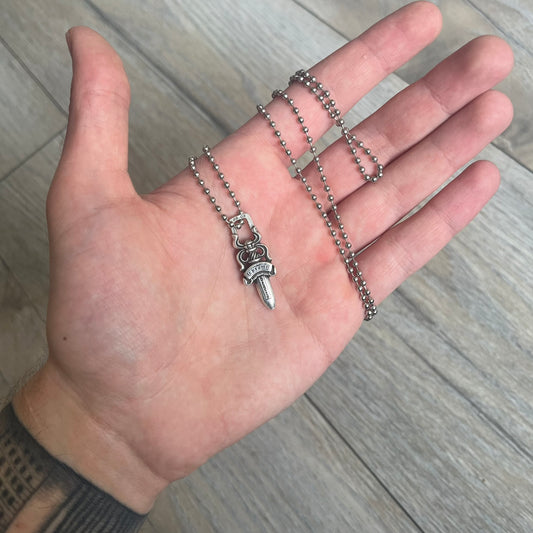 Chrome Hearts Dagger Zipper Pendant crafted from .925 silver, featuring the brand’s signature double-sided dagger design with “Chrome” on the guard and “Hearts” on the blade. Comes on a standard steel ball chain (non-CH). Square bail for chain attachment with 925 tag on one side and 89 03 on the other. The dagger was originally a zipper pull.