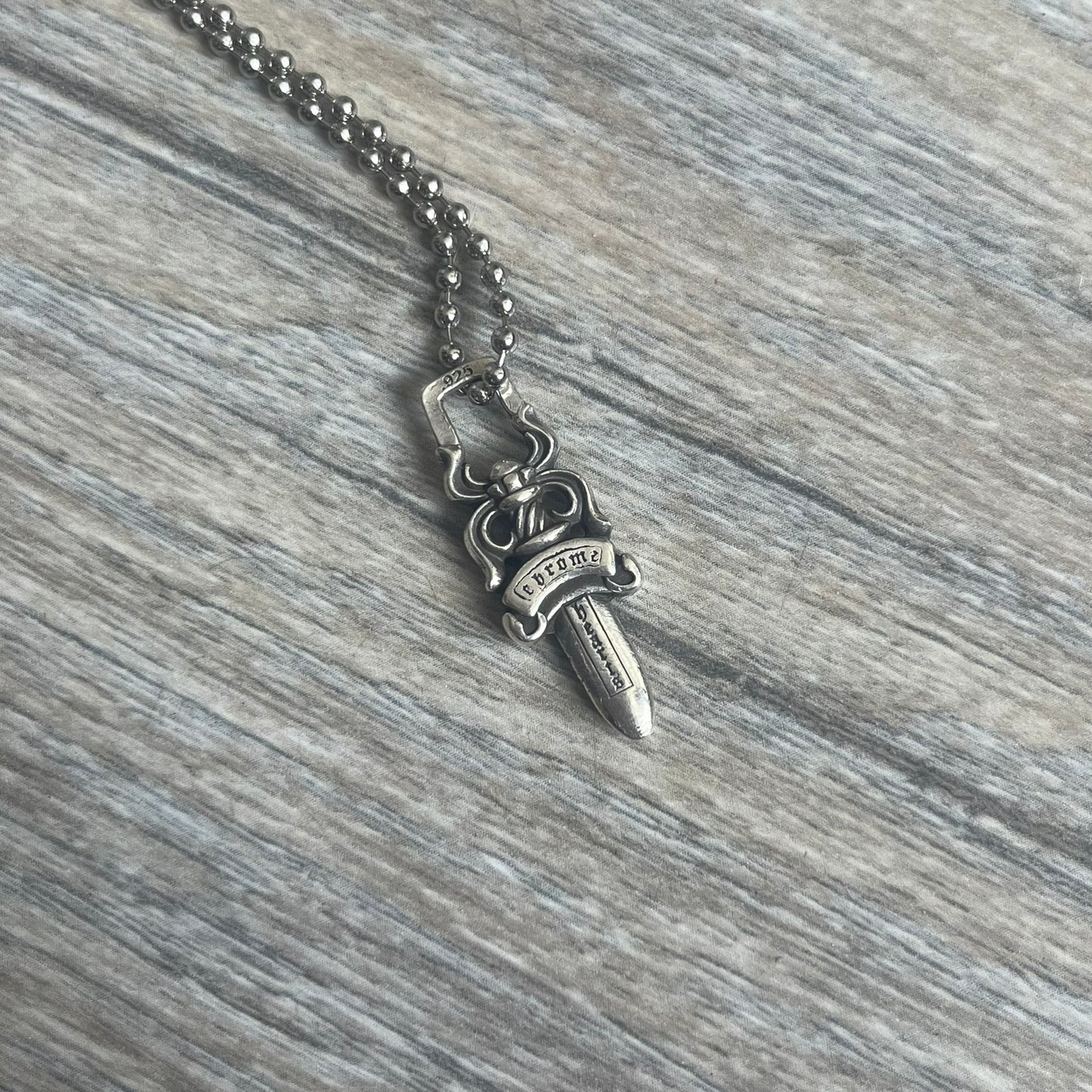 Chrome Hearts Dagger Zipper Pendant crafted from .925 silver, featuring the brand’s signature double-sided dagger design with “Chrome” on the guard and “Hearts” on the blade. Comes on a standard steel ball chain (non-CH). Square bail for chain attachment with 925 tag on one side and 89 03 on the other. The dagger was originally a zipper pull.