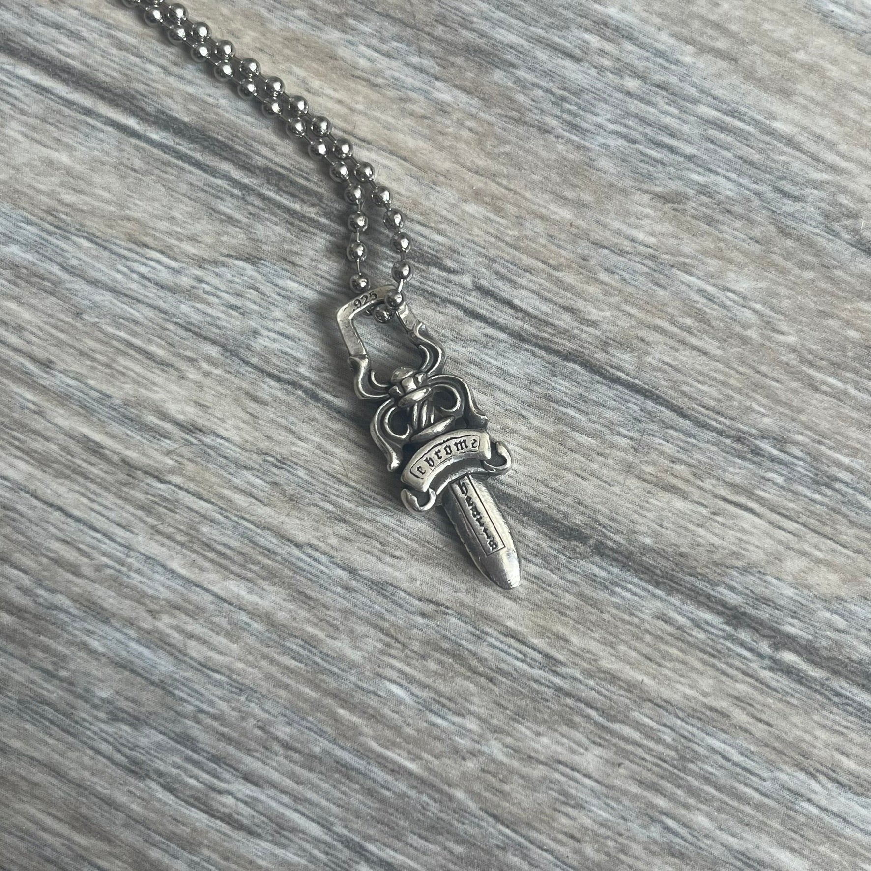 Chrome Hearts Dagger Zipper Pendant crafted from .925 silver, featuring the brand’s signature double-sided dagger design with “Chrome” on the guard and “Hearts” on the blade. Comes on a standard steel ball chain (non-CH). Square bail for chain attachment with 925 tag on one side and 89 03 on the other. The dagger was originally a zipper pull.