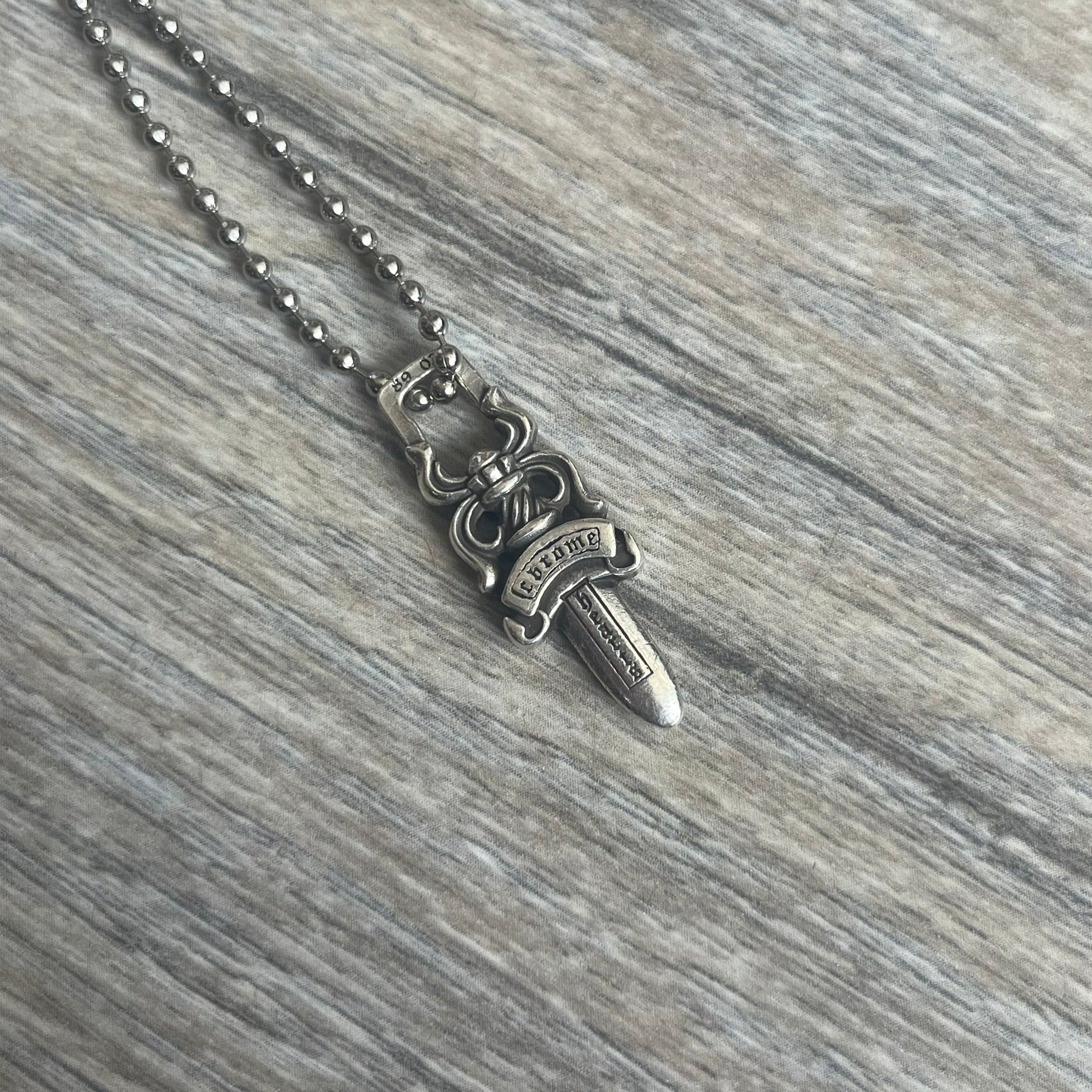 Chrome Hearts Dagger Zipper Pendant crafted from .925 silver, featuring the brand’s signature double-sided dagger design with “Chrome” on the guard and “Hearts” on the blade. Comes on a standard steel ball chain (non-CH). Square bail for chain attachment with 925 tag on one side and 89 03 on the other. The dagger was originally a zipper pull.