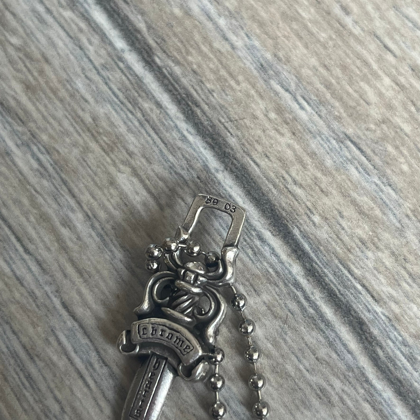 Chrome Hearts Dagger Zipper Pendant crafted from .925 silver, featuring the brand’s signature double-sided dagger design with “Chrome” on the guard and “Hearts” on the blade. Comes on a standard steel ball chain (non-CH). Square bail for chain attachment with 925 tag on one side and 89 03 on the other. The dagger was originally a zipper pull.