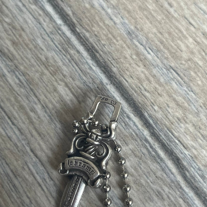 Chrome Hearts Dagger Zipper Pendant crafted from .925 silver, featuring the brand’s signature double-sided dagger design with “Chrome” on the guard and “Hearts” on the blade. Comes on a standard steel ball chain (non-CH). Square bail for chain attachment with 925 tag on one side and 89 03 on the other. The dagger was originally a zipper pull.