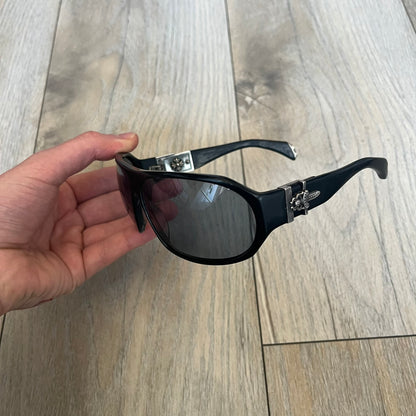 Chrome Hearts Hung Shield Black Sunglasses are black sunglasses from Chrome Hearts featuring black lenses and detailed metal hardware on the arms. The outer arm joints are finished with dagger emblems, while the inner sides feature Chrome Hearts cross motifs. The arm tips include metal end pieces with engraved Chrome Hearts branding. The pair comes with a black leather Chrome Hearts case.