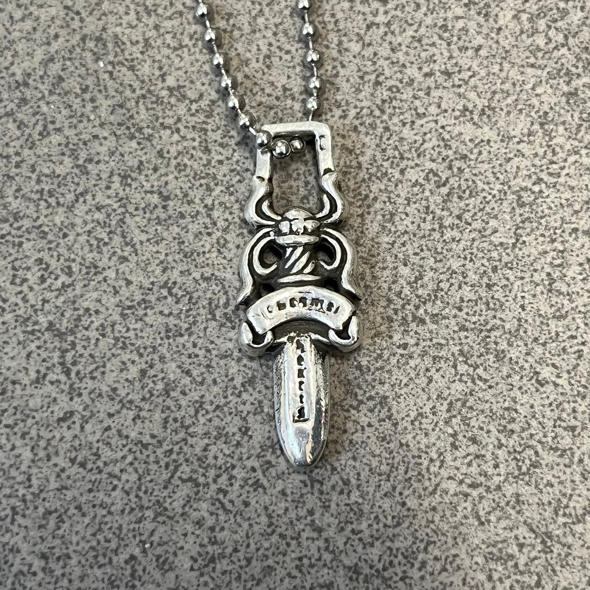 Chrome Hearts #5 Keychain Dagger Necklace crafted from .925 silver, featuring the brand’s signature double-sided dagger design with “Chrome” on the guard and “Hearts” on the blade. Comes on a stainless steel ball chain (non-CH). Square bail for chain attachment, the dagger was originally a keychain.