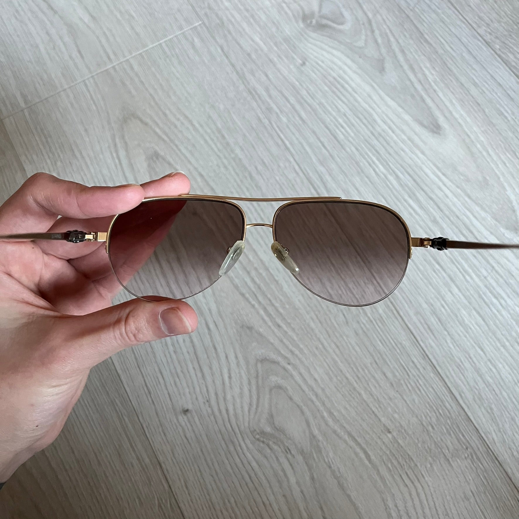 Chrome Hearts Stains Gold Sunglasses featuring a gold-tone metal frame with brown mirror-coated plastic lenses. Made in Japan, this model includes aged silver Chrome Hearts cross detailing wrapped around the arms, cut-out cross accents at the arm tips, and visible Chrome Hearts branding on the bridge and top of the frame. Additional cross branding appears on the nose pads. Comes complete with the original black leather Chrome Hearts zip case, finished with exterior branding and a dagger-shaped zipper pull.