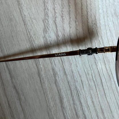 Chrome Hearts Stains Gold Sunglasses featuring a gold-tone metal frame with brown mirror-coated plastic lenses. Made in Japan, this model includes aged silver Chrome Hearts cross detailing wrapped around the arms, cut-out cross accents at the arm tips, and visible Chrome Hearts branding on the bridge and top of the frame. Additional cross branding appears on the nose pads. Comes complete with the original black leather Chrome Hearts zip case, finished with exterior branding and a dagger-shaped zipper pull.