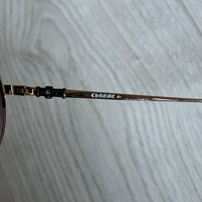 Chrome Hearts Stains Gold Sunglasses featuring a gold-tone metal frame with brown mirror-coated plastic lenses. Made in Japan, this model includes aged silver Chrome Hearts cross detailing wrapped around the arms, cut-out cross accents at the arm tips, and visible Chrome Hearts branding on the bridge and top of the frame. Additional cross branding appears on the nose pads. Comes complete with the original black leather Chrome Hearts zip case, finished with exterior branding and a dagger-shaped zipper pull.