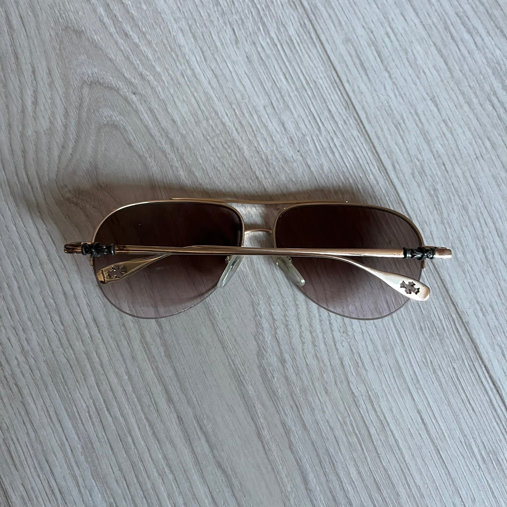 Chrome Hearts Stains Gold Sunglasses featuring a gold-tone metal frame with brown mirror-coated plastic lenses. Made in Japan, this model includes aged silver Chrome Hearts cross detailing wrapped around the arms, cut-out cross accents at the arm tips, and visible Chrome Hearts branding on the bridge and top of the frame. Additional cross branding appears on the nose pads. Comes complete with the original black leather Chrome Hearts zip case, finished with exterior branding and a dagger-shaped zipper pull.