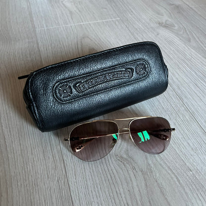 Chrome Hearts Stains Gold Sunglasses featuring a gold-tone metal frame with brown mirror-coated plastic lenses. Made in Japan, this model includes aged silver Chrome Hearts cross detailing wrapped around the arms, cut-out cross accents at the arm tips, and visible Chrome Hearts branding on the bridge and top of the frame. Additional cross branding appears on the nose pads. Comes complete with the original black leather Chrome Hearts zip case, finished with exterior branding and a dagger-shaped zipper pull.