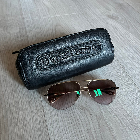 Chrome Hearts Stains Gold Sunglasses featuring a gold-tone metal frame with brown mirror-coated plastic lenses. Made in Japan, this model includes aged silver Chrome Hearts cross detailing wrapped around the arms, cut-out cross accents at the arm tips, and visible Chrome Hearts branding on the bridge and top of the frame. Additional cross branding appears on the nose pads. Comes complete with the original black leather Chrome Hearts zip case, finished with exterior branding and a dagger-shaped zipper pull.