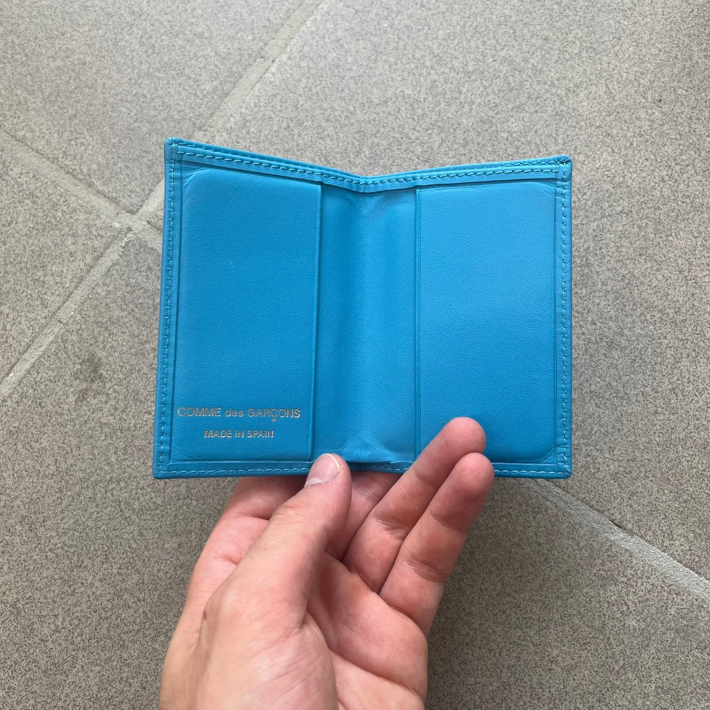Comme des Garçons Blue Bifold Leather Wallet features a clean and minimal bi-fold design crafted from smooth aqua blue calf leather. The interior includes multiple card slots, a banknote compartment, and a gold-tone Comme des Garçons logo stamp for a subtle branded detail. A timeless everyday accessory combining practical organization with the brand’s signature minimalist aesthetic.

