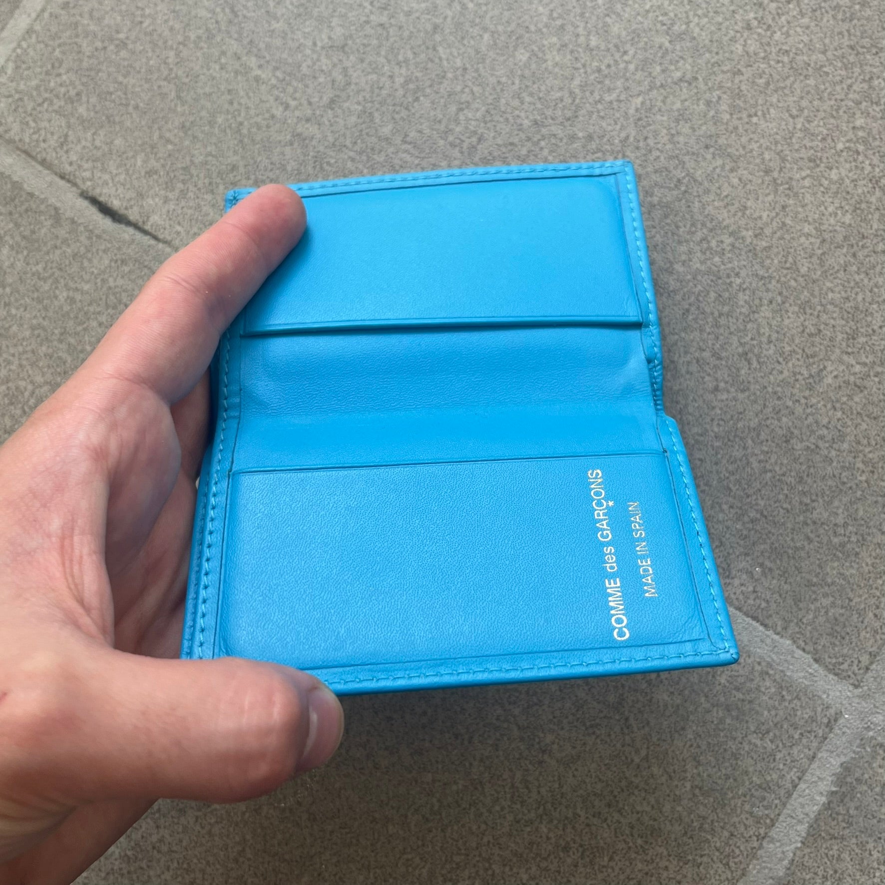 Comme des Garçons Blue Bifold Leather Wallet features a clean and minimal bi-fold design crafted from smooth aqua blue calf leather. The interior includes multiple card slots, a banknote compartment, and a gold-tone Comme des Garçons logo stamp for a subtle branded detail. A timeless everyday accessory combining practical organization with the brand’s signature minimalist aesthetic.
