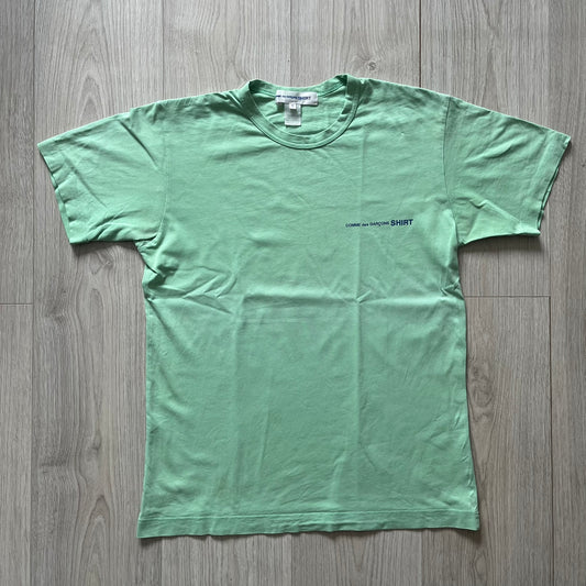 Green t-shirt with a small logo on a wooden floor
