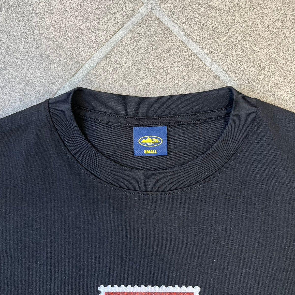 Corteiz x Skepta Konnichiwa 10th Anniversary BlackTee is a 100% cotton T-shirt released to mark the 10th anniversary of Skepta's "Konnichiwa" album, featuring the original album cover artwork with a red postage stamp graphic of Skepta. The design includes a "First Class" ink stamp alongside the Corteiz logo replacing the original cover's "London" mark.