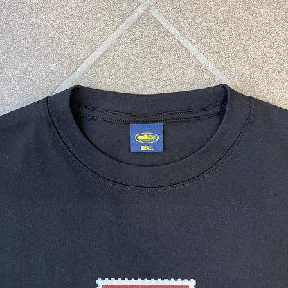 Corteiz x Skepta Konnichiwa 10th Anniversary BlackTee is a 100% cotton T-shirt released to mark the 10th anniversary of Skepta's "Konnichiwa" album, featuring the original album cover artwork with a red postage stamp graphic of Skepta. The design includes a "First Class" ink stamp alongside the Corteiz logo replacing the original cover's "London" mark.
