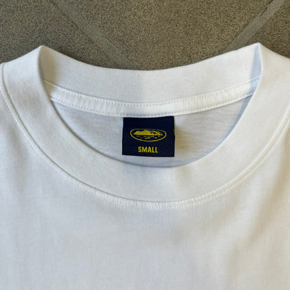Corteiz x Skepta Konnichiwa 10th Anniversary White Tee is a 100% cotton T-shirt released to mark the 10th anniversary of Skepta's "Konnichiwa" album, featuring the original album cover artwork with a red postage stamp graphic of Skepta. The design includes a "First Class" ink stamp alongside the Corteiz logo replacing the original cover's "London" mark.