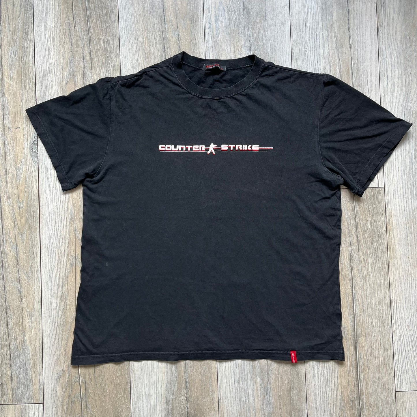 Counter-Strike 2000s Logo Tee is a black T-shirt featuring the classic Counter-Strike logo printed across the front. The design includes white “Counter” and “Strike” text separated by the armed character silhouette, with a red shadow layer behind the logo. A small red Ben Hao tag is attached near the bottom hem.
