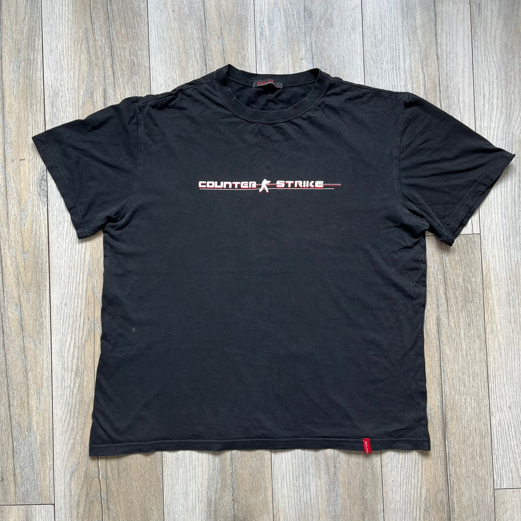 Counter-Strike 2000s Logo Tee is a black T-shirt featuring the classic Counter-Strike logo printed across the front. The design includes white “Counter” and “Strike” text separated by the armed character silhouette, with a red shadow layer behind the logo. A small red Ben Hao tag is attached near the bottom hem.