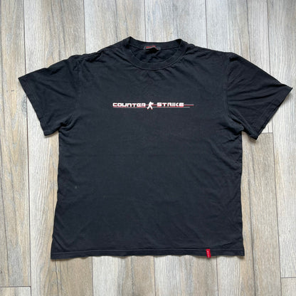 Counter-Strike 2000s Logo Tee is a black T-shirt featuring the classic Counter-Strike logo printed across the front. The design includes white “Counter” and “Strike” text separated by the armed character silhouette, with a red shadow layer behind the logo. A small red Ben Hao tag is attached near the bottom hem.